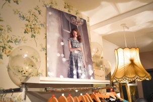 A photograph of a large Emily and Rose marketing print sitting on top of a clothing rail with pretty floral wallpaper decorating the wall behind and two large lampshades either side