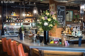 A photograph of the left side of the bar with silver bucket of prosecco bottles and a large vase of flowers. Photo by Tom Gold.